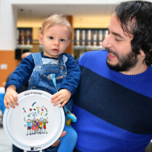 A father is holding his child in his arms and smiling at it. The child is holding an empty children's plate.